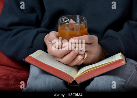 Senior kaukasischen Mann, der Becher mit Heißgetränk mit beiden Händen ruht auf Buch hält. Gemütlich zu halten, während des Lesens Konzept Stockfoto