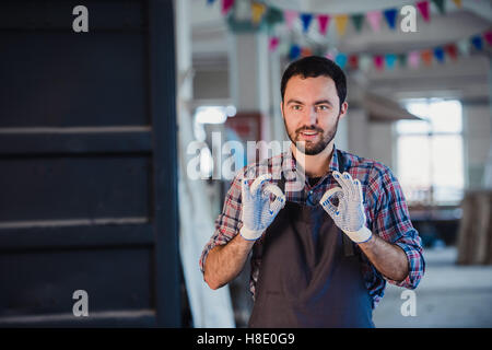 Lächelnde Arbeiter gestikulieren Doppel OK Schild am workshop Stockfoto