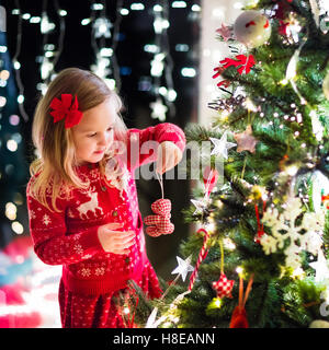 Kleines Mädchen in rot Strickpullover nordische Rentier hängende Ornamente auf Weihnachtsbaum mit Licht, Tand und Candy Canes. Stockfoto