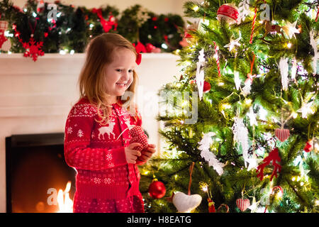 Kleines Mädchen in rot Strickpullover nordische Rentier hängende Ornamente auf Weihnachtsbaum mit Licht, Tand und Candy Canes. Stockfoto