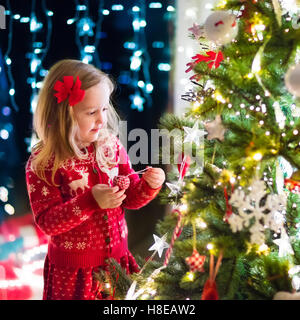 Kleines Mädchen in rot Strickpullover nordische Rentier hängende Ornamente auf Weihnachtsbaum mit Licht, Tand und Candy Canes. Stockfoto