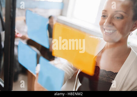 Lächelnde junge Frau kleben Kleber Geschäftsnotizen an Glaswand. Glaswand aufsetzen Ideen. Stockfoto