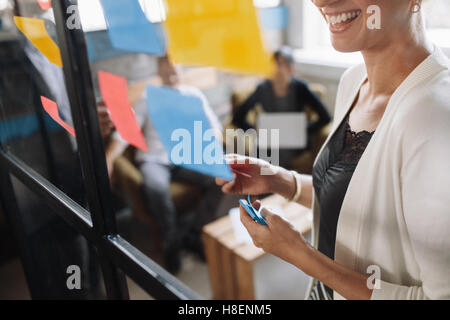 Lächelnde junge Frau brainstorming mit Klebstoff Notizen auf Glaswand. Kolleginnen und Kollegen vorlegen Ideen während der Sitzung. Stockfoto