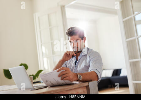 Aufnahme des jungen Mann mit Laptop am Tisch sitzen und Lesen von Dokumenten. Gut aussehend Geschäftsmann vom home-Office arbeiten. Stockfoto