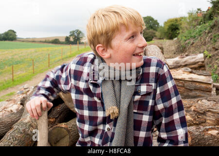 Junge im karierten Hemd, Schal und Mütze hält eine Axt vor einem Haufen Brennholz auf einem Bauernhof Stockfoto