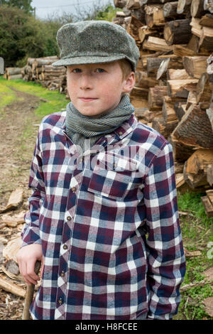 Junge im karierten Hemd, Schal und Mütze hält eine Axt vor einem Haufen Brennholz auf einem Bauernhof Stockfoto