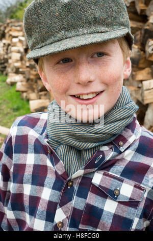 Junge im karierten Hemd, Schal und Mütze hält eine Axt vor einem Haufen Brennholz auf einem Bauernhof Stockfoto