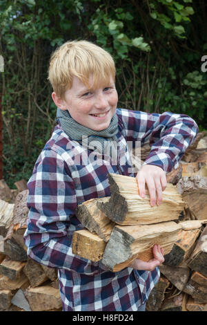 Junge im karierten Hemd, Schal und Mütze hält eine Axt vor einem Haufen Brennholz auf einem Bauernhof Stockfoto