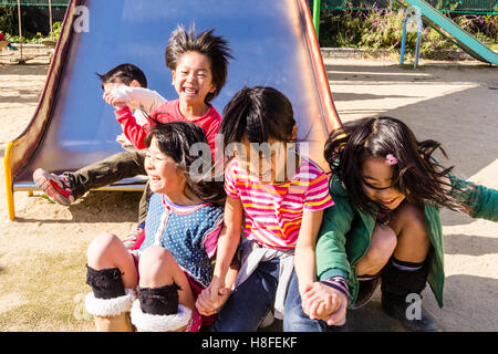 Japan. Schule Spielplatz. Kinder, drei Mädchen, 6-8 Jahre alt, springen Sie in der Zeile, aufgeregt und glücklich zu schreien. Zwei Jungen Rutschen hinter Ihnen Stockfoto