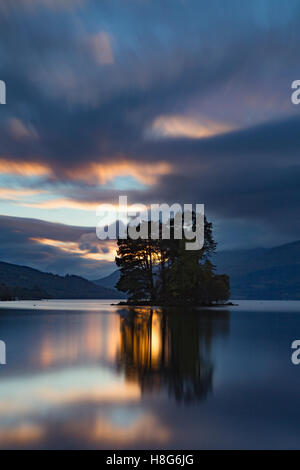 Loch Tay, Perthshire, Schottland, reflektiert das Licht von der untergehenden Sonne, wie es geht hinter den Bäumen auf einer der Inseln. Stockfoto