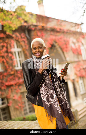 Moderne junge afroamerikanische Frau posiert mit Kaffee zum mitnehmen und Outdoor-Handy Stockfoto