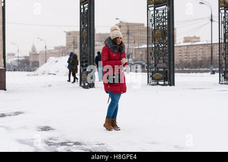 Moskau, Russland. Samstag, 12. November 2016. Frühwinter in Moskau. Keine Sonne, bewölkt. Leichte frost bis zu-5 Grad Celsius (+ 23F) durch den Abend, stetige Schneefall, aber nicht sehr schwer. Unbekannte schöne junge Frau im roten Mantel und blaue Jeans wartet, bis jemand durch den Eingang zum Gorky Park. Bildnachweis: Alex Bilder/Alamy Live-Nachrichten Stockfoto