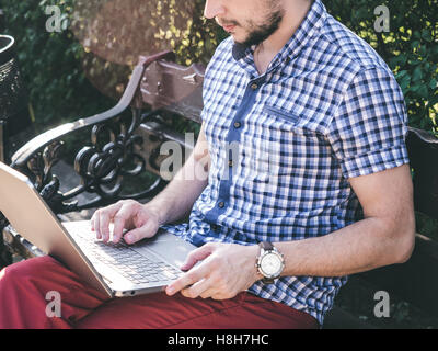 Junger Mann arbeitet an seinem Laptop sitzt auf der Bank im Park, modernes Design notebook Stockfoto