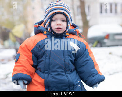 Attraktiven jungen spielen mit dem ersten Schnee. Er lächelt und schaut Schneemann. Dick blau-Orange Overall hell gestreiften Hut auf ein Jahr altes Kind. Stockfoto