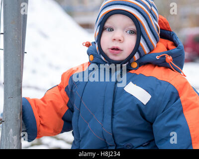 Attraktiven jungen spielen mit dem ersten Schnee. Er lächelt und schaut Schneemann. Dick blau-Orange Overall hell gestreiften Hut auf ein Jahr altes Kind. Stockfoto