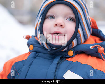 Attraktiven jungen spielen mit dem ersten Schnee. Er lächelt und schaut Schneemann. Dick blau-Orange Overall hell gestreiften Hut auf ein Jahr altes Kind. Stockfoto