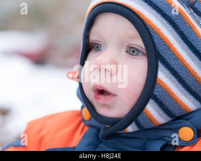 Attraktiven jungen spielen mit dem ersten Schnee. Er lächelt und schaut Schneemann. Dick blau-Orange Overall hell gestreiften Hut auf ein Jahr altes Kind. Stockfoto