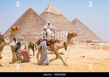 Kairo, Ägypten-Touristen und Kameltreiber mit ihren Kamelen zu Fuß durch die Wüste mit den drei großen Pyramiden von Gizeh in th Stockfoto