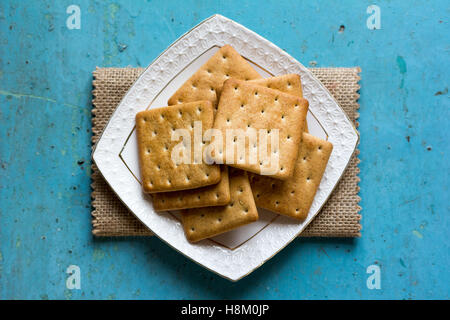 Cracker-Kekse in eine weiße Keramik quadratische Untertasse auf Leinen Serviette auf alten blauen Hintergrund Nahaufnahme. Ansicht von oben Stockfoto