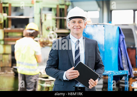 Porträt von zuversichtlich reifer Geschäftsmann hält digitale Tablet mit Arbeiter im Hintergrund im Werk Stockfoto