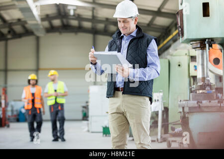 Ältere männliche Inspektor schreiben in Zwischenablage beim Arbeitnehmer im Hintergrund im Werk Stockfoto