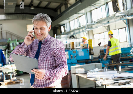 Reifen männlichen Vorgesetzten Blick auf Zwischenablage beim Telefonieren mit Handy in der Industrie Stockfoto