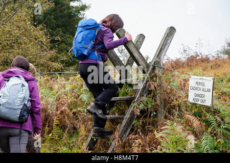 Zweisprachige "Gefahr fernzuhalten" Zeichen auf einem öffentlichen Wanderweg bei Wanderern einen Leiter-Stil über einen Stacheldrahtzaun zu klettern. Wales UK Stockfoto