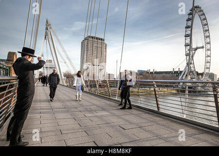 Ein jüdischer Junge trägt einen traditionellen schwarzen Hut mit breiter Krempe fotografiert zwei Mädchen auf Hungerford Bridge, London Stockfoto