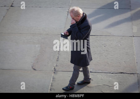 Von oben betrachtet, hält ein Mann eine Objektiv auf ein Auge, um sein Handy zu lesen, als er einen Platz geht. Stockfoto