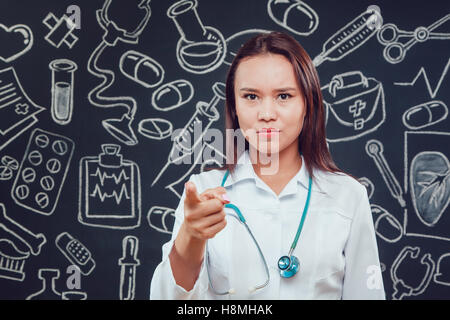 Junge Ärztin im weißen Kittel stehen auf dunklem Hintergrund mit Muster. Er zeigt mit dem Finger nach vorne Stockfoto