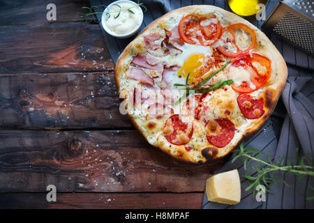 Pizza Carbonara mit Speck, Salami, Parmesan-Käse und Huhn-Ei am rustikalen Holztisch Stockfoto