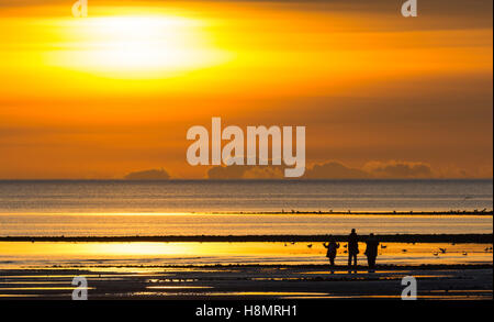 Sonnenuntergang an einem bewölkten Tag an der Küste mit einer kleinen Familie in Silhouette beobachten. Stockfoto