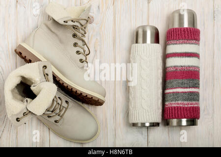 Leichte Winterstiefel und eine Thermoskanne in den gestrickten Deckel auf dem hellen Holz Hintergrund. Stockfoto