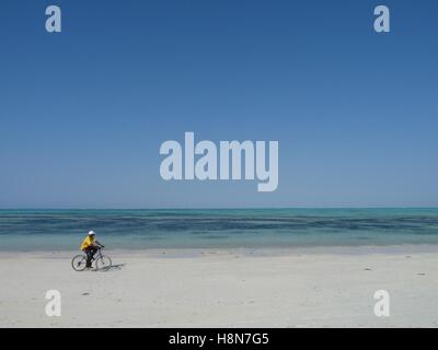 Ein junger Mann fährt ein Fahrrad entlang einer leeren weißen Sandstrand mit türkisblauem Meer und strahlend blauen Himmel im Hintergrund Stockfoto