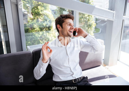 Fröhliche entspannte Jungunternehmer zeigen Ordnung Geste und reden über Handy im Büro Stockfoto