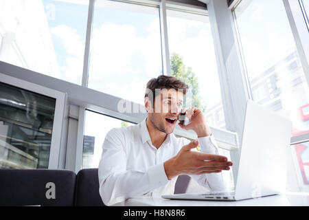 Erstaunt junger Geschäftsmann mit Laptop und Handy telefonieren im Büro Stockfoto