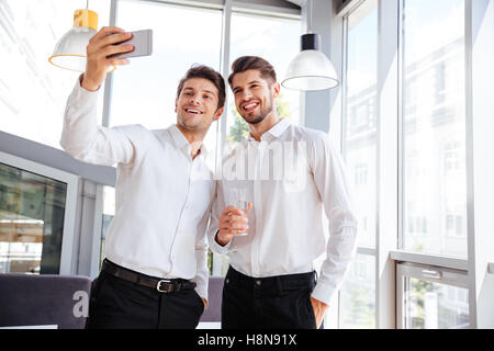 Zwei fröhliche junge Geschäftsleute stehen und nehmen Selfie im Büro Stockfoto