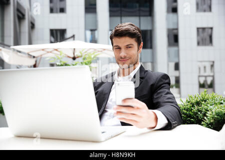 Lächelnd jungen Geschäftsmann mit Laptop sitzen und mit Handy im Café im freien Stockfoto