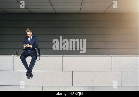 Junger Geschäftsmann auf schlichte weiße Mauer sitzend und mit Telefon Stockfoto