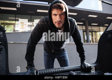 Kriminelle junger Mann in schwarzer Hoodie und Handschuhe stehen und blickte im Kofferraum eines Autos Stockfoto