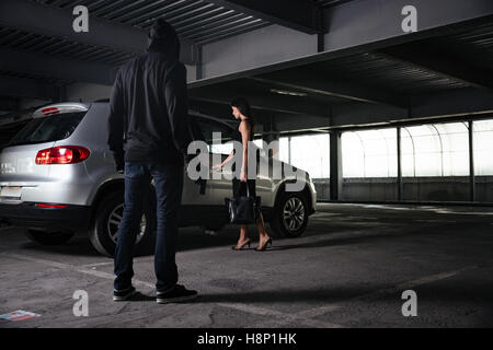 Kriminelle Mann in schwarzer Hoodie stehen und blickte auf junge Frau Eröffnung Auto auf dem Parkplatz Stockfoto