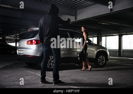 Mann-Dieb in schwarzer Hoodie stehend und drohte mit Pistole, junge Frau auf Parkplatz Stockfoto