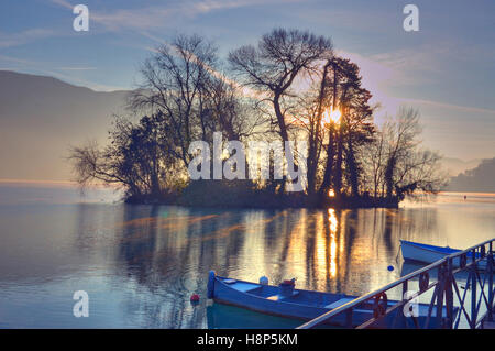 Morgenlicht durch die Bäume auf einer kleinen Insel im See von Annecy Frankreich mit kleinen Fischerbooten gefesselt im Vordergrund. Stockfoto