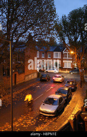 Herbst Blätter bedecken die Bürgersteige und Straßen in einer typischen Süd-London-Straße nach Einbruch der Dunkelheit auf eine nasse Nacht. Ein Radfahrer geht. Stockfoto