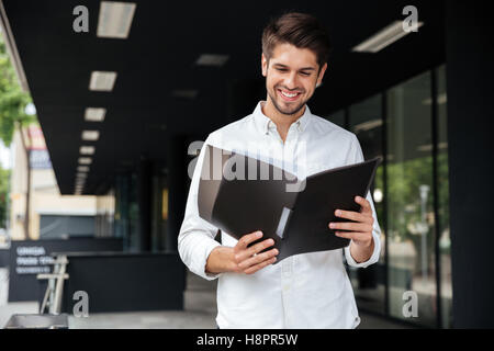 Glücklich hübscher junger Geschäftsmann stehen und blickte durch Dokumente im Ordner "" Stockfoto