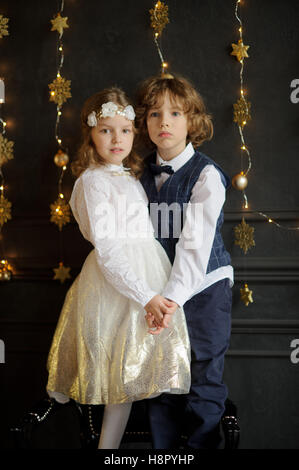 Zwei bezaubernde Kinder stehen Hände beigetreten. Das Mädchen ist in einem weißen und goldenen elegante Kleid gekleidet. Auf den Kopf von ihr eine Wre Stockfoto