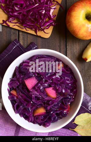 Geschmorter Rotkohl mit Apfel in Schale umgeben von Zutaten, overhead auf dunklem Holz mit Tageslicht fotografiert Stockfoto