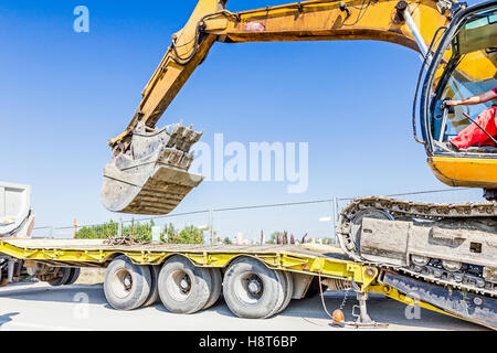 Schwere Bagger klettert es auf niedrige Plattform Anhänger über rückseitige Rampe, trägt zwei Eimer ineinander eingefügt. Stockfoto