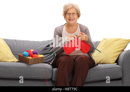 Reife Frau sitzt auf einem Sofa stricken und schaut in die Kamera, die isoliert auf weißem Hintergrund Stockfoto