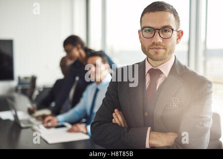 Schöne bärtige Professional in Brillen und Krawatte stehen mit verschränkten Armen neben drei beschäftigten Kollegen vor großen Offi